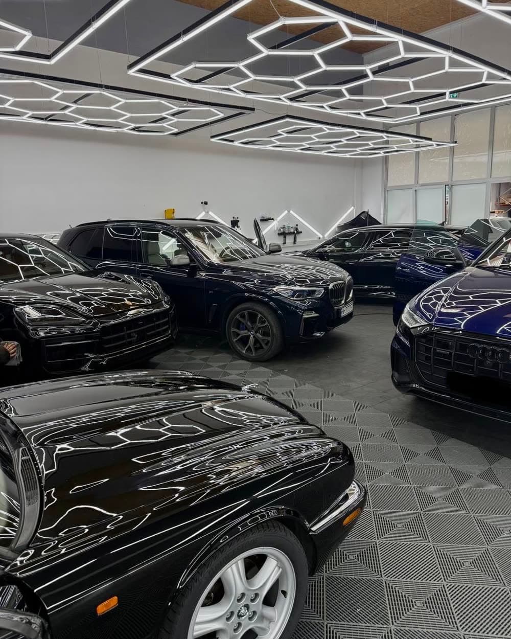 Mehrere Fahrzeuge nebeneinander in der Halle, darunter ein dunkelblauer BMW und ein Audi; in jedem Lack stehen die Waben der Deckenleuchten.
