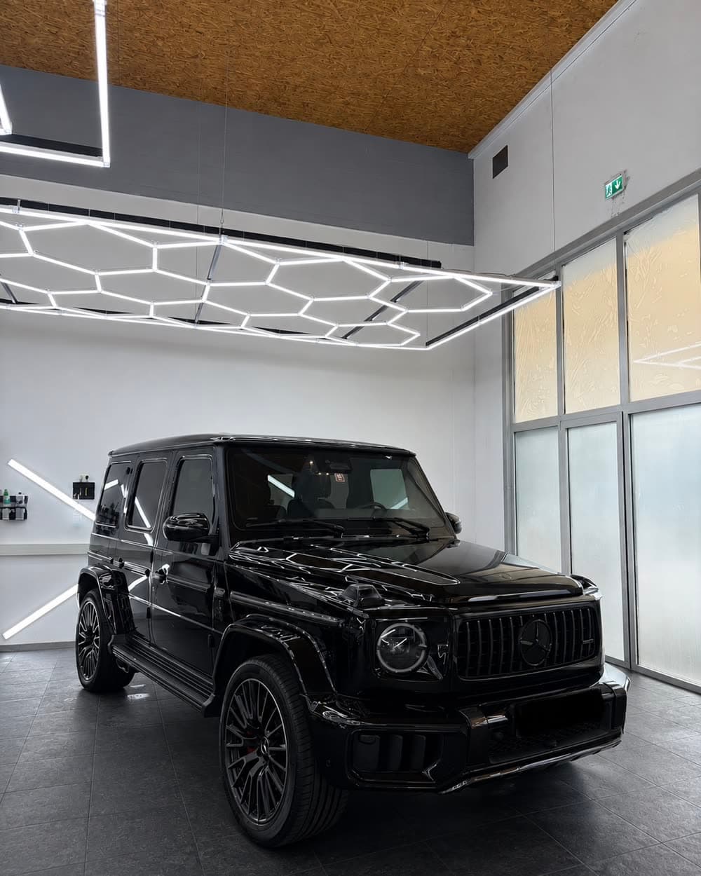 Schwarze Mercedes G-Klasse schräg von vorne in der hellen Halle, dahinter die Glasfront; die Wabenleuchten der Decke liegen als Linien auf Haube und Kotflügel.